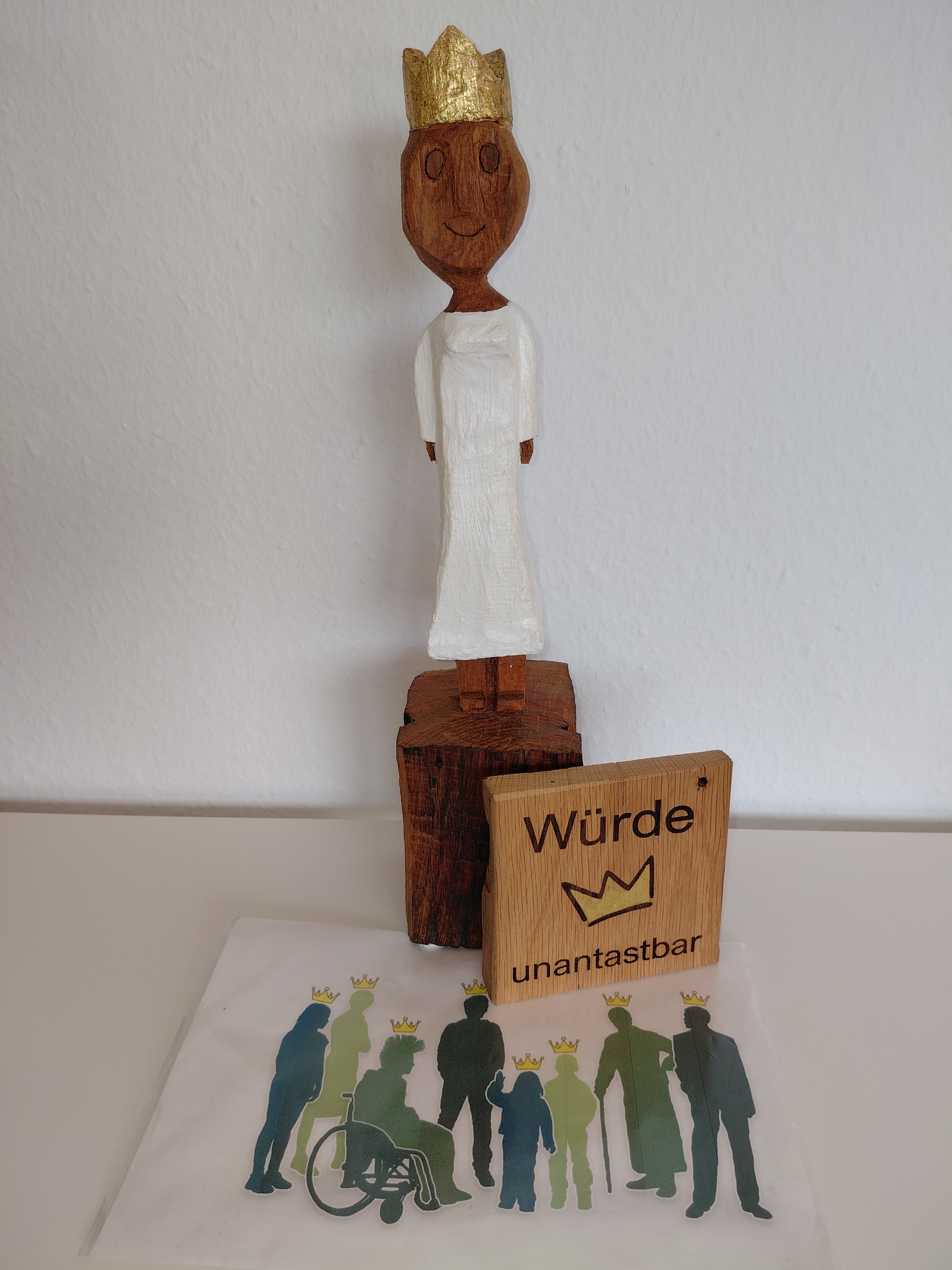 Königin - Würde unantastbar Eine aus Holz geschnitzte Königin. Davor steht eine Holztafel mit einer goldenen Krone und den eingebrannten Wörtern 'Würde unantastbar'. Außerdem noch eine Grafik mit verschiedenen Menschen, die alle die gleiche goldenen Krone tragen